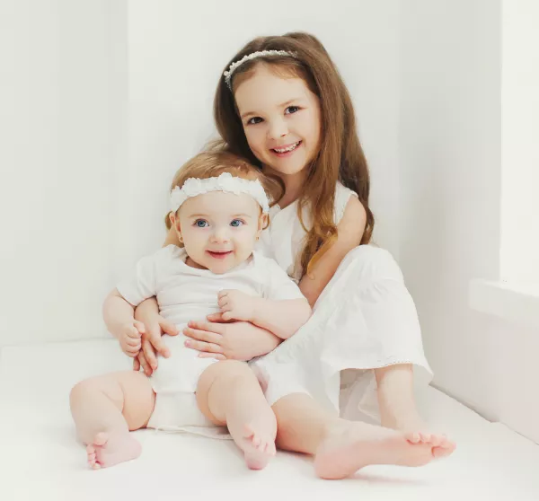 Portrait Of Cute Little Girl Child With Her Sister Baby Sitting In White Room At Home Near Window